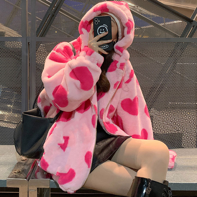 Frau in rosa Plüschjacke mit Herzmuster, hält Smartphone, sitzt auf Bank, modisch, urban.