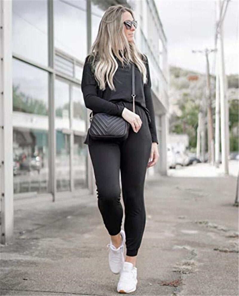 Frau in schwarzem Sportanzug, Sonnenbrille, weiße Sneaker, schwarze Umhängetasche, urbaner Hintergrund.