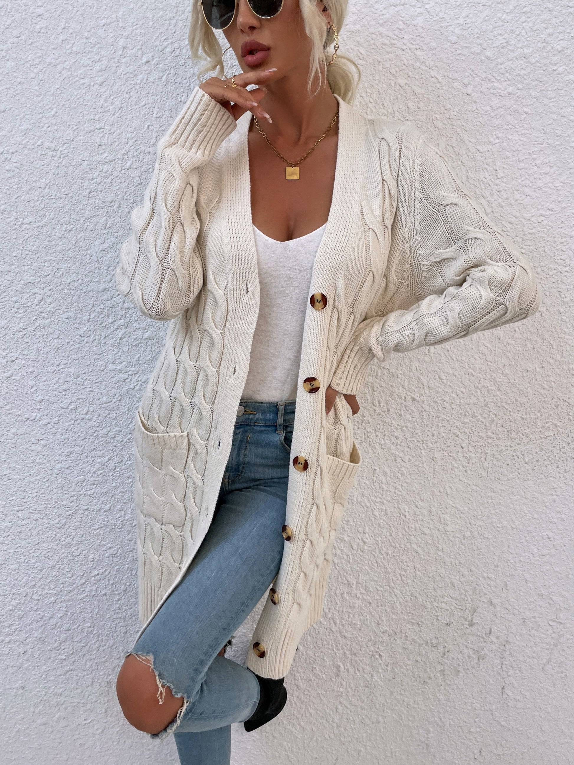 Frau in cremefarbenem Strickcardigan mit Knöpfen, weiße Bluse, zerrissene Jeans, Sonnenbrille.