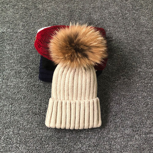 Beige Strickmütze mit Kunstfellbommel auf grauem Hintergrund, Wintermode Accessoire.