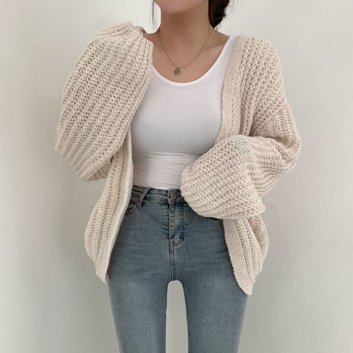 Frau in cremefarbenem Strickcardigan, weißem Top und blauer Jeans vor weißem Hintergrund.