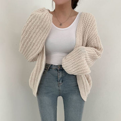 Frau in cremefarbenem Strickcardigan, weißem Top und blauer Jeans vor weißem Hintergrund.