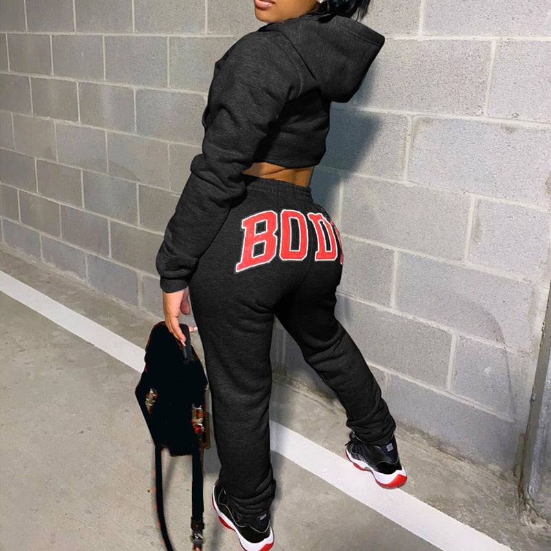 Frau in schwarzem Hoodie und Jogginghose mit "BODY"-Aufdruck, Turnschuhe, graue Wand.
