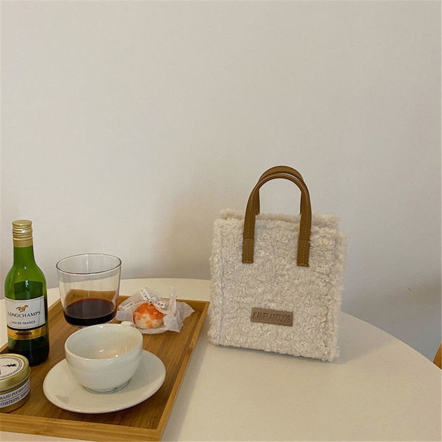 Weiße Plüschhandtasche mit braunen Griffen auf Tisch neben Tablett mit Getränken und Snacks.