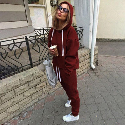 Frau in rotem Hoodie-Set mit Kapuze, hält Kaffeebecher, trägt Sonnenbrille und weiße Sneaker.