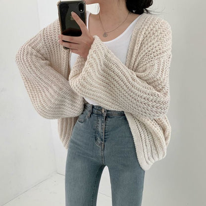Frau in cremefarbenem Strickcardigan, weißem T-Shirt und blauer Jeans, Smartphone in der Hand.