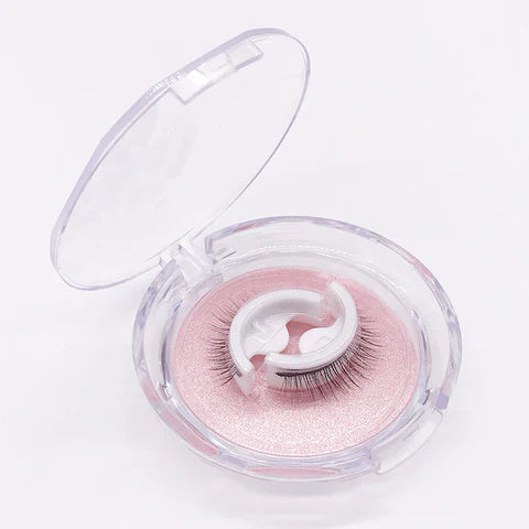 Ett par lösögonfransar på vit hållare ligger i en öppen, rund genomskinlig förpackning med rosa glittrande botten. 1