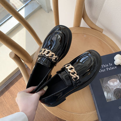 Schwarze Lackleder-Loafer mit goldener Kette auf Holzstuhl, modische Damenschuhe.