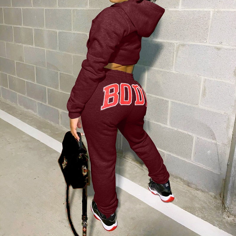 Frau in bordeauxfarbenem Jogginganzug mit "BODY"-Aufdruck, schwarze Sneaker, graue Wand.