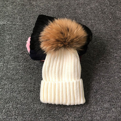 Weiße Strickmütze mit braunem Kunstfellbommel auf grauem Hintergrund.