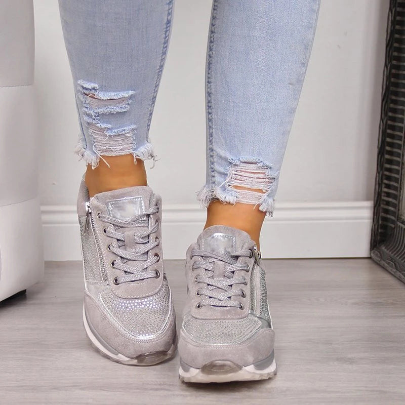Framifrån av gråa sneakers på fötterna, med tät snörning, dragkedjor på ytterkanterna och glittrande stenar på tåpartiet. 5