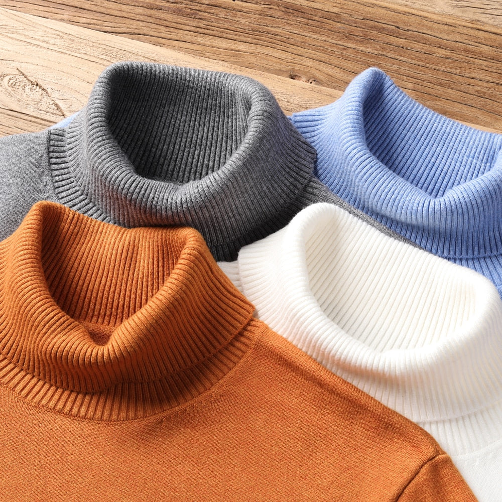 Vier Rollkragenpullover aus Strick in Grau, Blau, Weiß und Orange auf Holztisch.