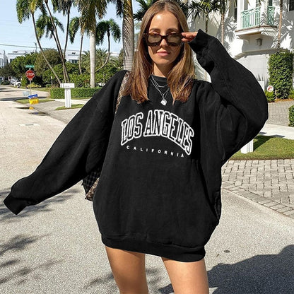 Frau in schwarzem "Los Angeles" Oversize-Pullover, Sonnenbrille, Straße, Palmen im Hintergrund.