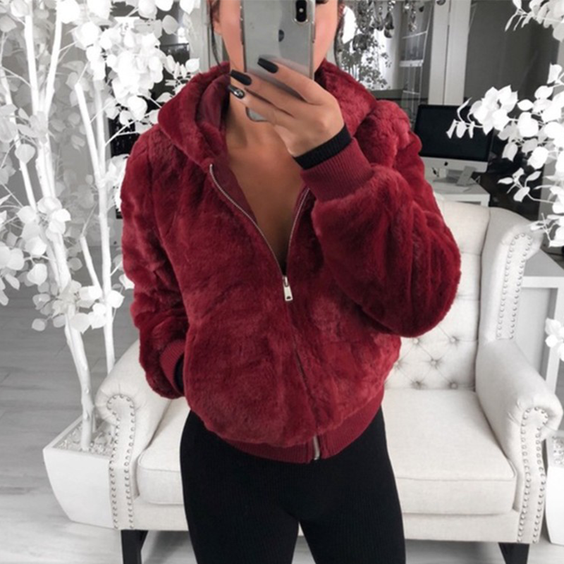 Frau in roter Kunstpelzjacke mit Kapuze, stehend vor weißem Sofa, modisch, gemütlich.