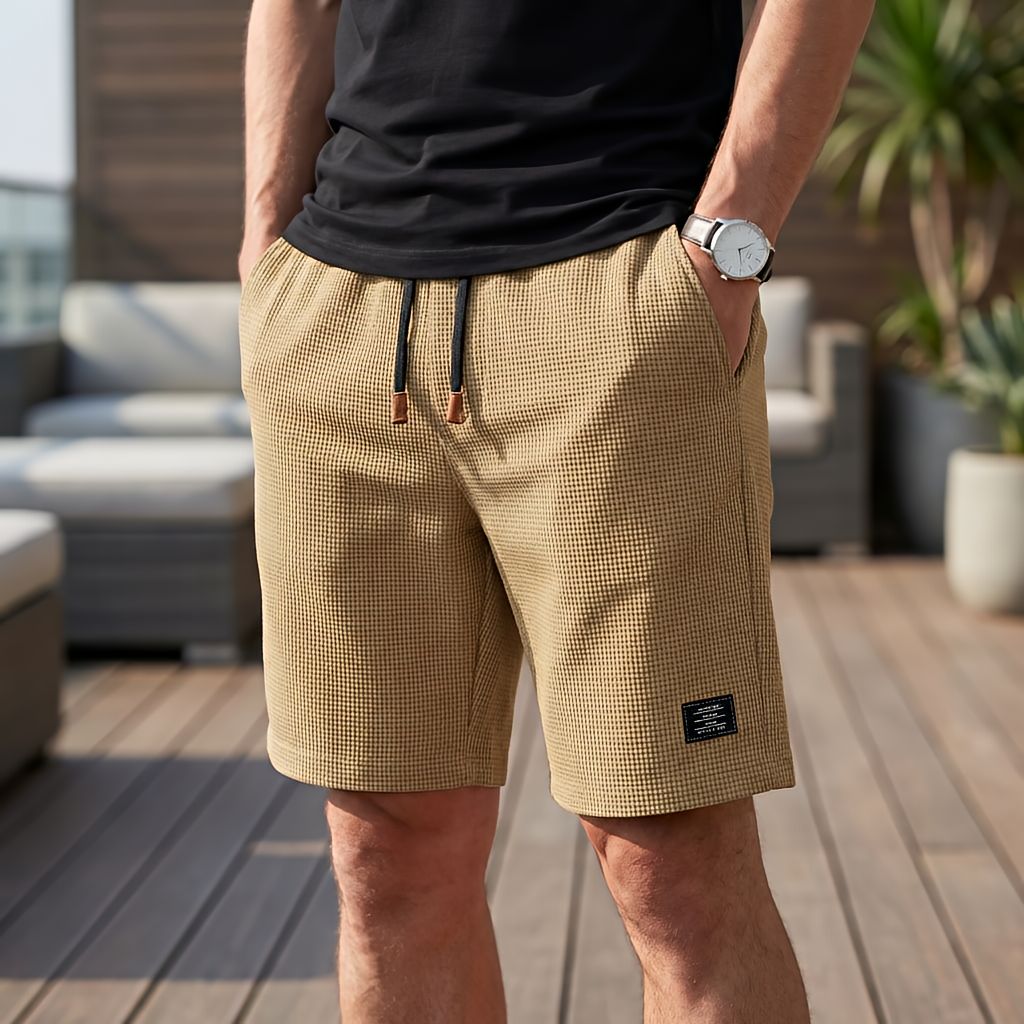 Sandfärgade shorts i vaffeltyg med svart dragsko och brun ändhylsa, burna med handen i fickan på en terrass. 4