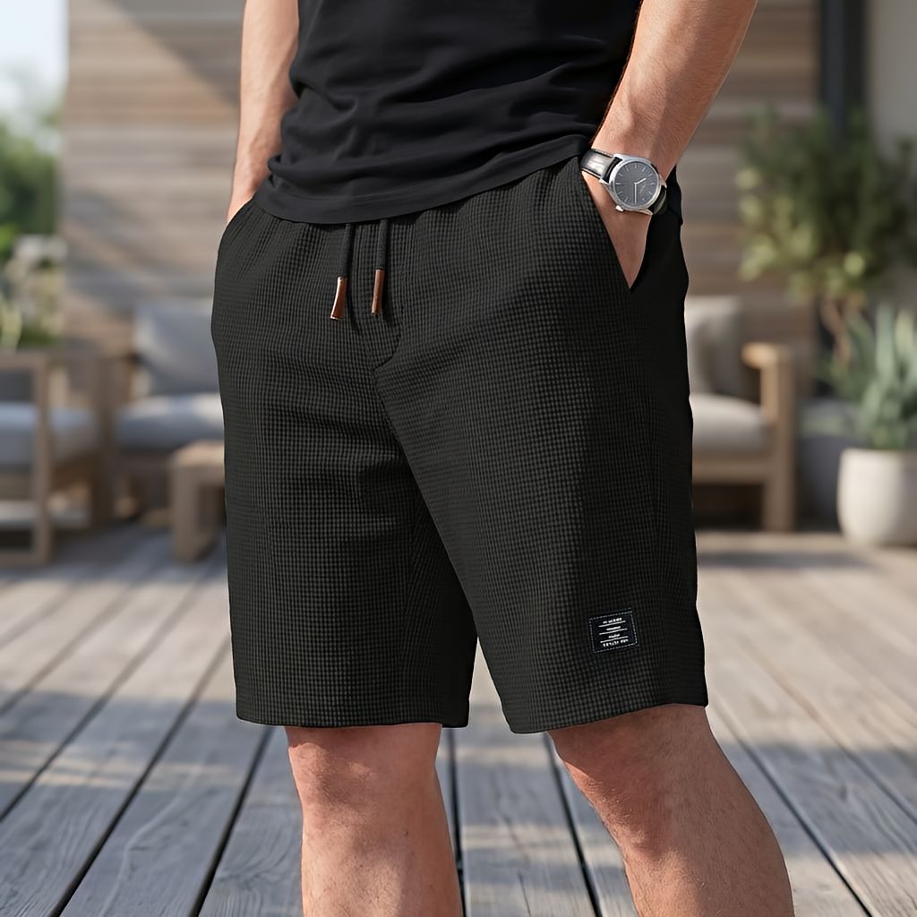 Svarta shorts i vaffeltyg med dragsko, burna av en man i svart t-shirt med armbandsur på en trädäck. 3