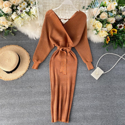 Braunes Strickkleid mit Gürtel, V-Ausschnitt, langen Ärmeln, neben Strohhut und Handtasche.