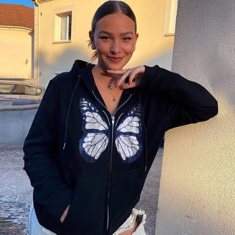 Frau in schwarzem Hoodie mit Schmetterlingsmotiv, lächelnd, draußen bei Sonnenuntergang.
