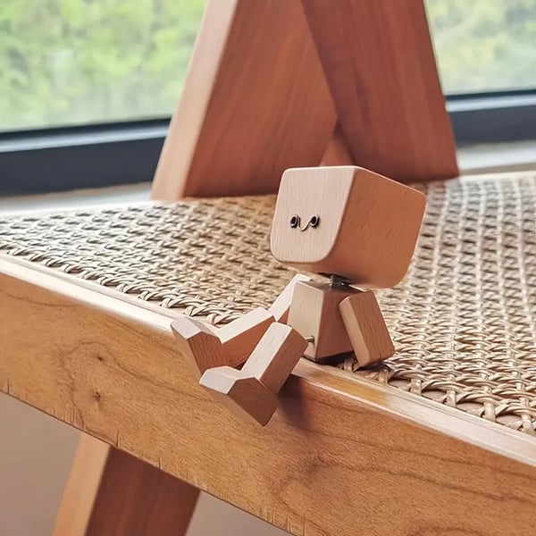 Liten träfigur med leende ansikte sittande på kanten av en stol med flätad sits av rotting. 5