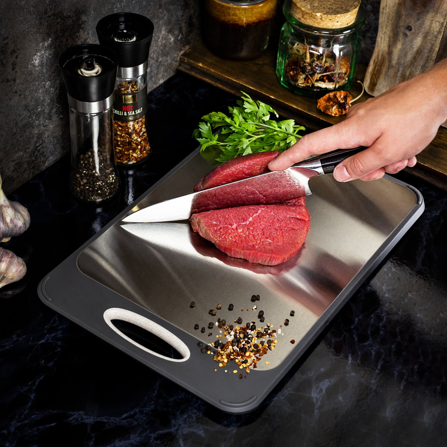 Hand schneidet rohes Rindfleisch auf Edelstahl-Schneidebrett, Gewürze und Kräuter im Hintergrund.
