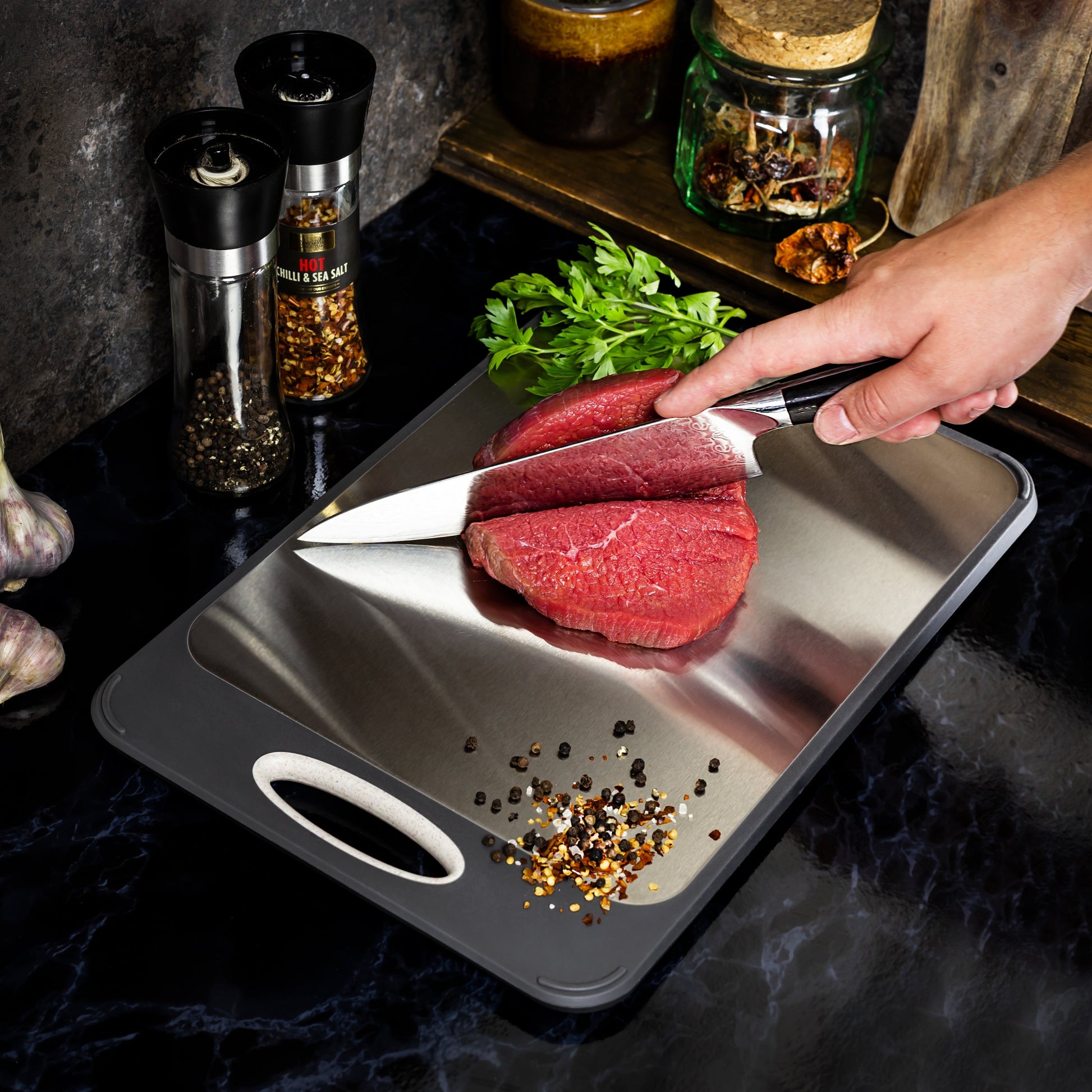 Hand schneidet rohes Rindfleisch auf Edelstahl-Schneidebrett, Gewürze und Kräuter im Hintergrund.