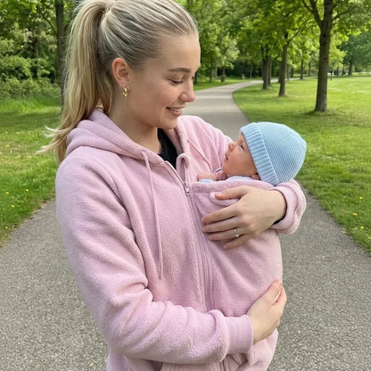 Kvinna i rosa fleecehoodie håller en bebis med blå mössa mot bröstet på en gångväg i en grön park. 4
