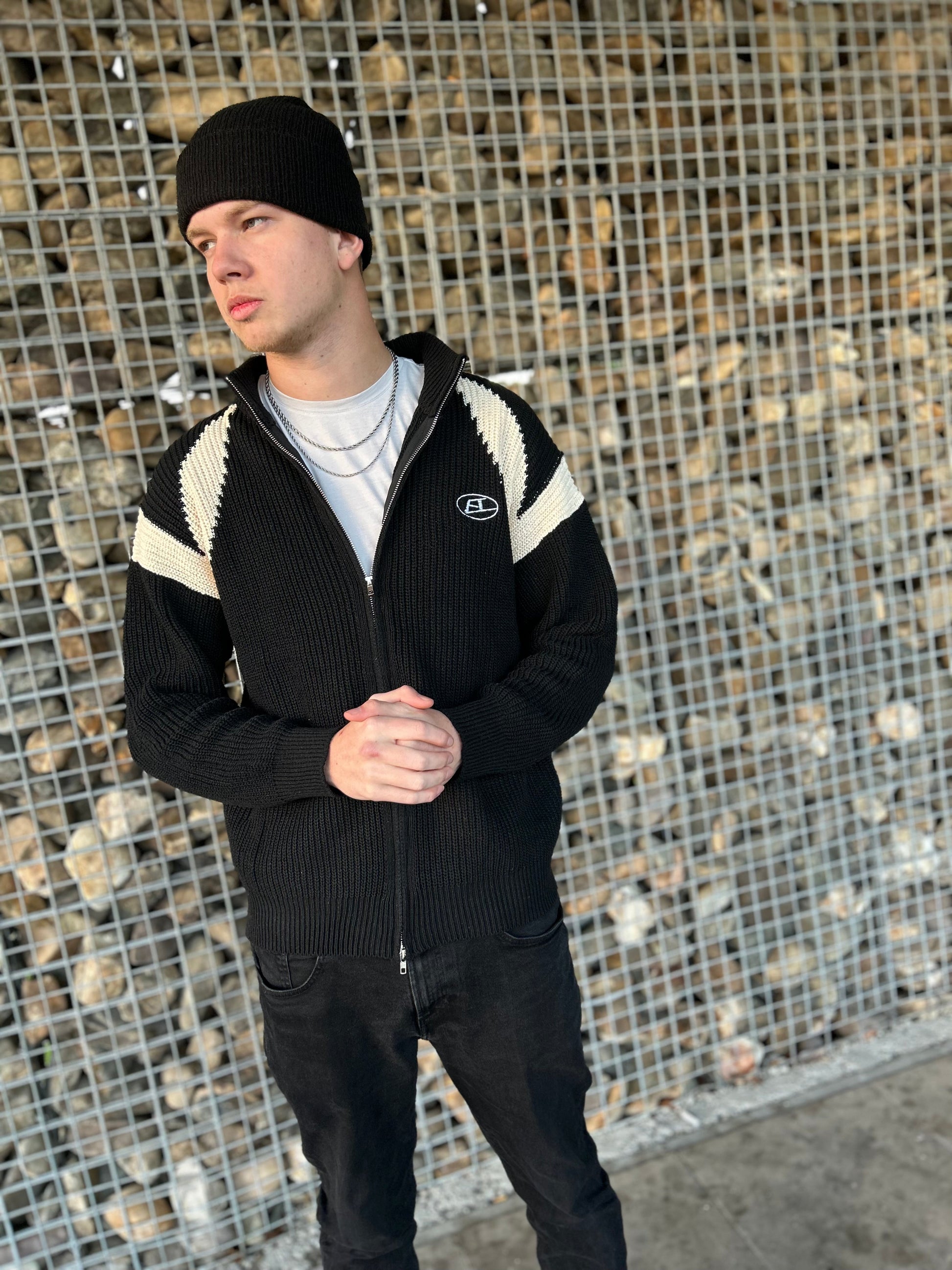 Mann in schwarzer Strickjacke mit weißen Streifen und Mütze vor Steinwand.