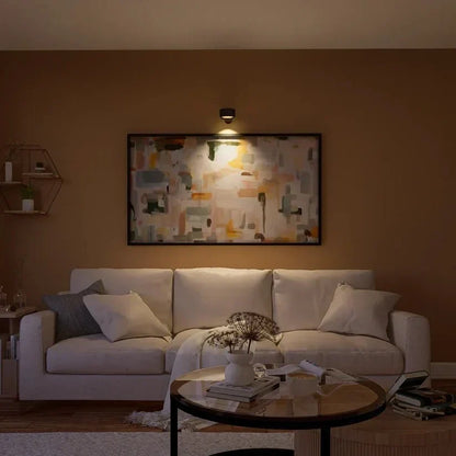Svart vägglampa lyser upp en målad tavla ovanför en ljus soffa i ett vardagsrum 15