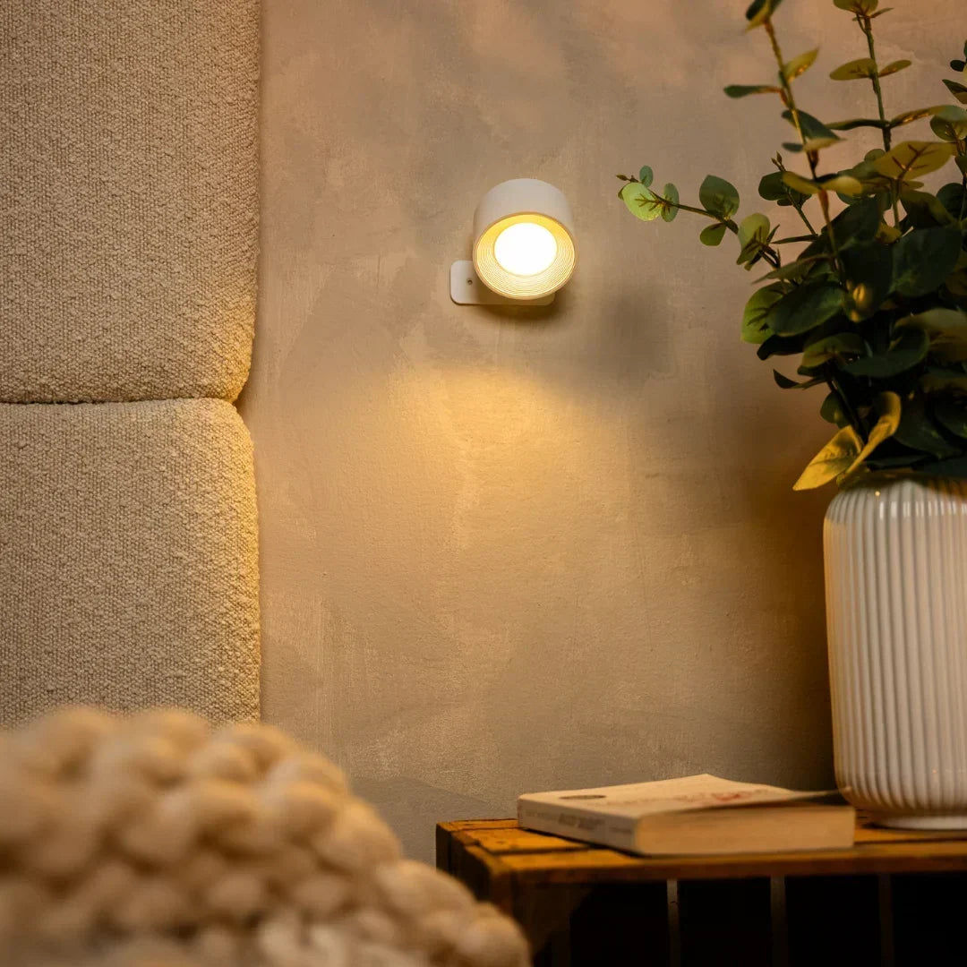 Vit vägglampa lyser varmt ovanför en träkrage med bok och en vas med grenar 7