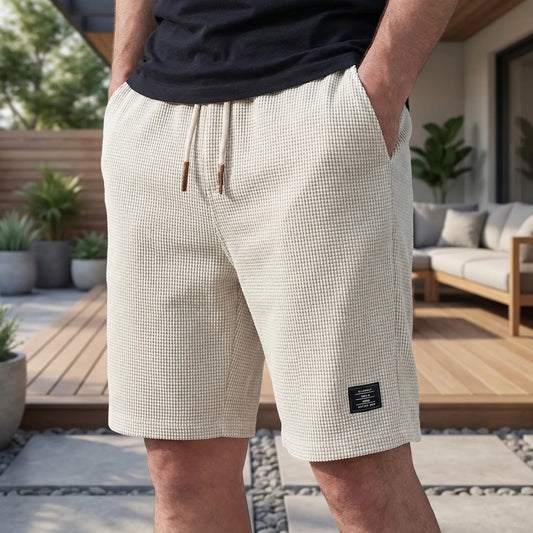 Närbild på shorts i ljust vaffelmönster med dragsko och små lädertoppar, burna med händerna i fickorna på en terrass. 2
