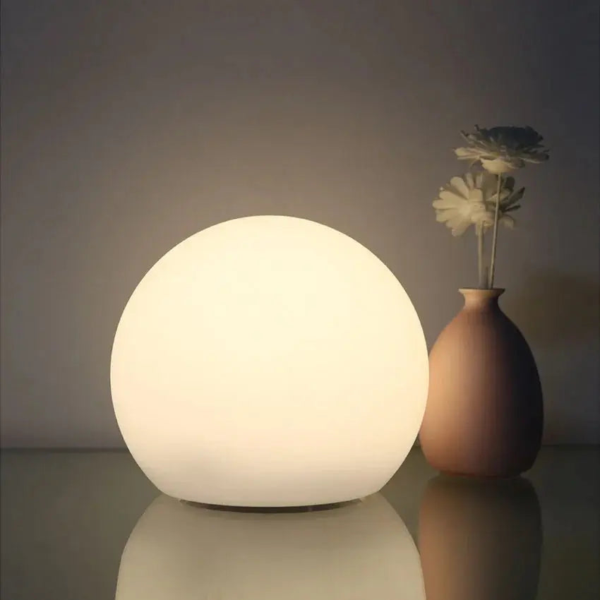 Tänd vit klotlampa med mjukt sken på en blank yta, bredvid en beige vas med två vita blommor. 2
