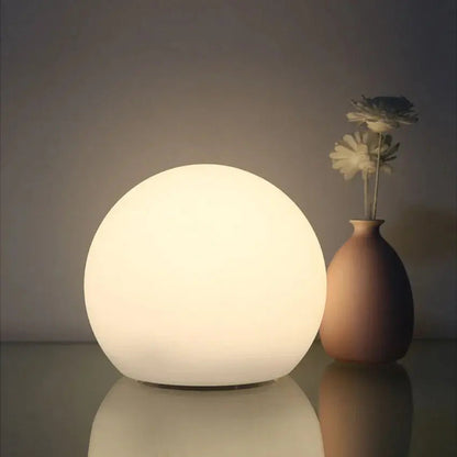 Tänd vit klotlampa med mjukt sken på en blank yta, bredvid en beige vas med två vita blommor. 2
