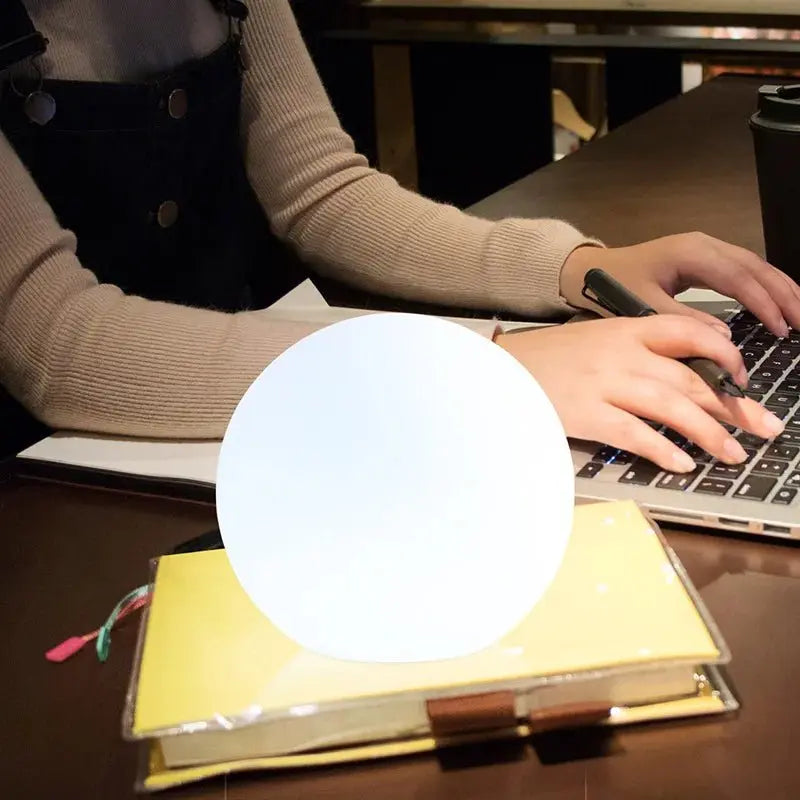 Klotlampa med vitt ljus placerad på en gul anteckningsbok vid ett skrivbord, medan en person i beige tröja skriver på en laptop. 7