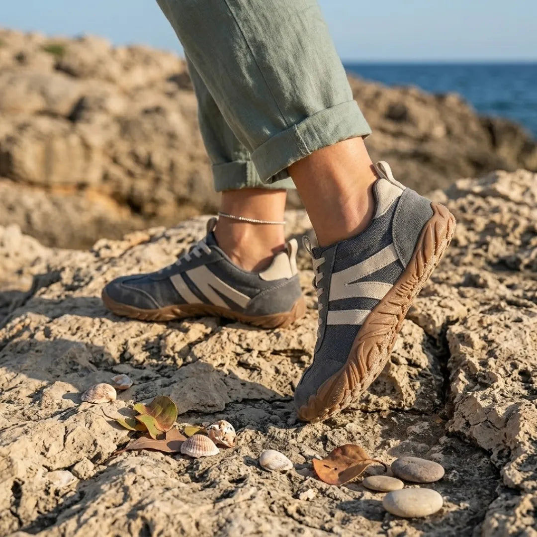 Grå skor med beige ränder och brun sula bärs av en person i gröna byxor på klippor vid havet. 2