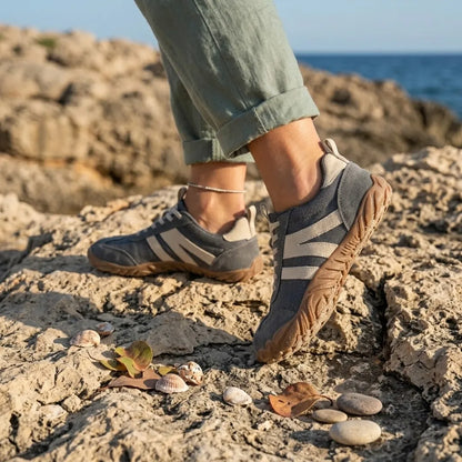 Grå skor med beige ränder och brun sula bärs av en person i gröna byxor på klippor vid havet. 2