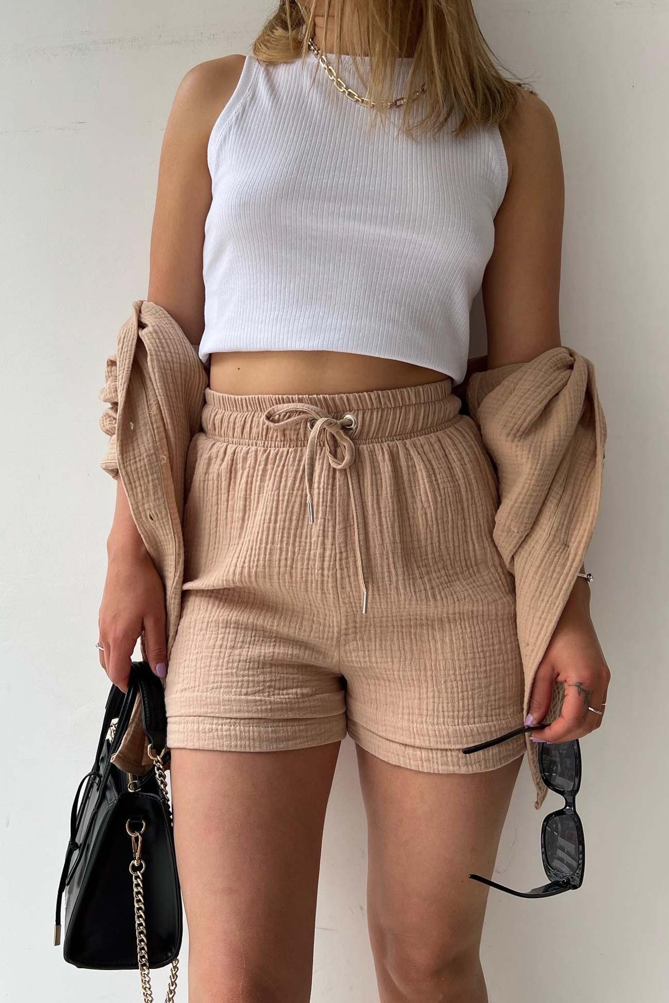 Frau in beigem Shorts-Set mit weißem Tanktop, hält schwarze Tasche und Sonnenbrille.