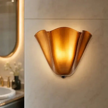 Tänd bärnstensfärgad glasvägglampa med vågig kant på marmorerad badrumsvägg, med spegel och handfat i bakgrunden. 10