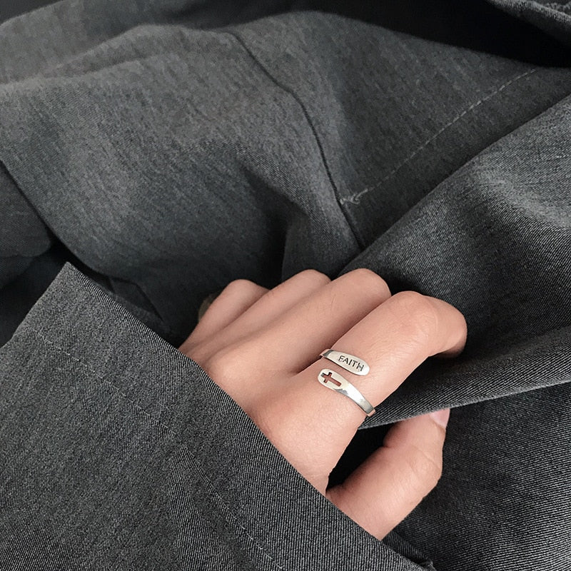 Silberner Ring mit Gravur "FAITH" und Kreuz, getragen an Hand, grauer Stoffhintergrund.