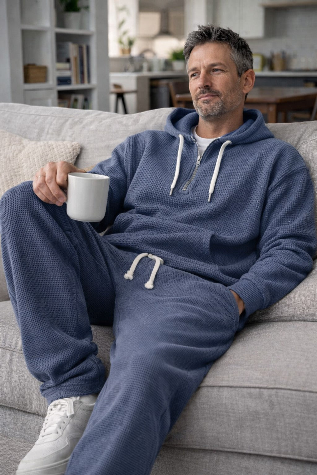 Man i blått våffelstickat set med huvtröja och joggingbyxor sitter avslappnat i en soffa och håller en vit kopp. 4