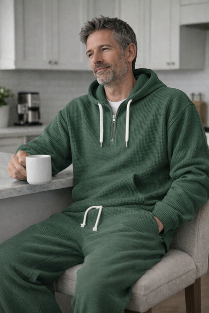 Man i grönt våffelstickat set sitter vid ett köksbord med en vit kaffekopp, med handen i huvtröjans ficka. 3