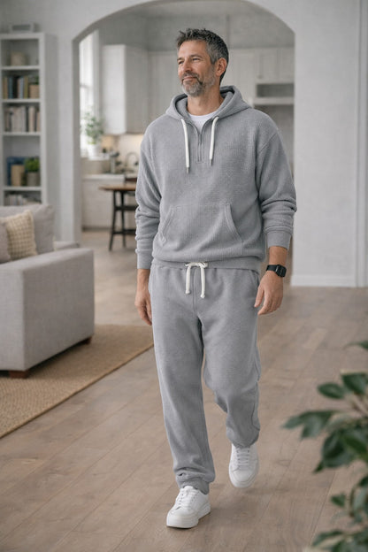 Man i grått våffelstickat set med huvtröja och joggingbyxor går genom ett ljust vardagsrum med valv, soffa och bokhylla. 2