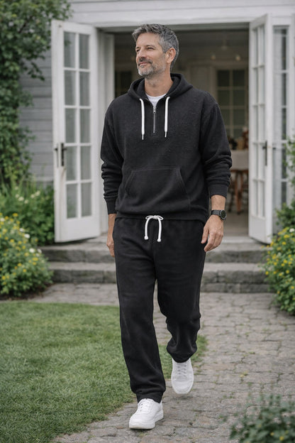 Man i svart våffelstickat set med huvtröja och joggingbyxor går på en stenlagd gång framför ett vitt hus med öppna dörrar. 1