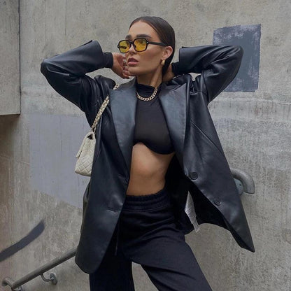 Frau in schwarzer Lederjacke, gelbe Sonnenbrille, schwarze Hose, weiße Tasche, urbane Umgebung.