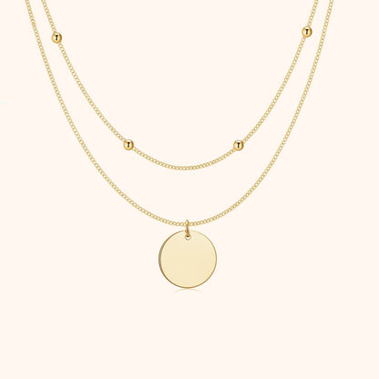 Zweireihige goldene Halskette mit rundem Anhänger und kleinen Buchstabenperlen.
