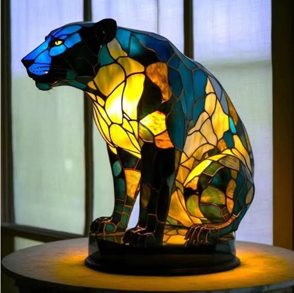 Lysande panterlampa i blå, grön och gul mosaik, sittande på en rund bas framför ett fönster. 16