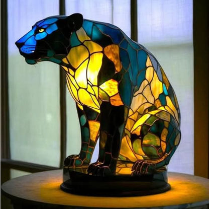 Lysande panterlampa i blå, grön och gul mosaik, sittande på en rund bas framför ett fönster. 16
