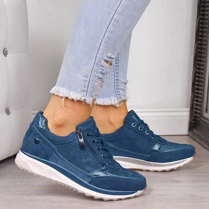 Blå sneakers med blixtlås vid snörningen och vit sula, bärda av en modell i slitna jeans på ljust golv. 5