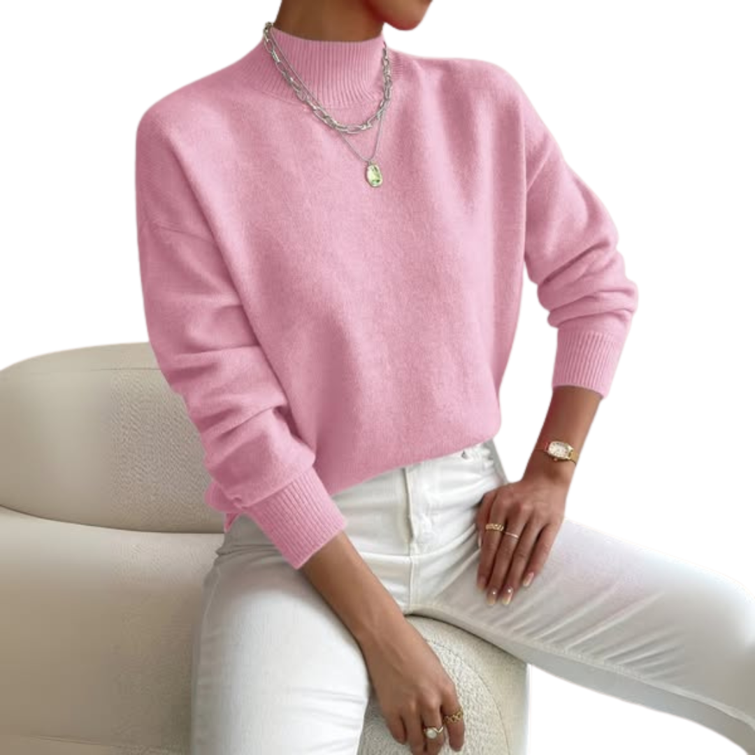 Modell i rosa lågpolo med ribbad krage och muddar, vita jeans och lager på lager-halsband, sittande på en vit fåtölj. 1
