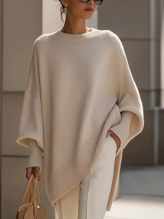 Kvinna bär den beigea stickade tröjan med ribbad asymmetrisk nederkant, vita byxor och en beige handväska, helfigur framifrån. 1