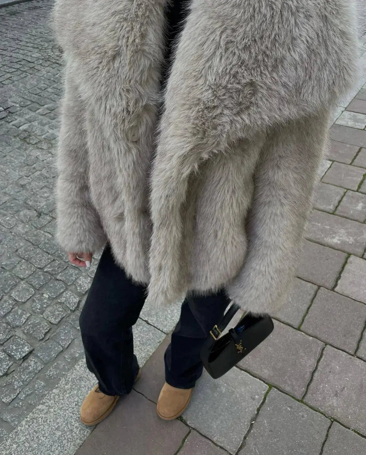 Grauer Kunstpelzmantel, schwarze Handtasche, braune Stiefel auf gepflastertem Gehweg.
